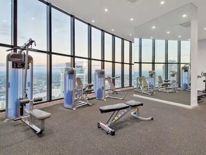 A gym with large windows, exercise equipment, and a view of the city. - Lvl50 Ocean Views | 3BR Skyhome | Pool+Gym+Parking (Surfers Paradise)