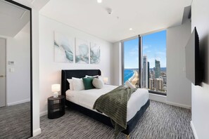 A modern hotel room with a large bed, a nightstand, a TV, and a view of the cityscape. - Lvl 50 Skyhome 3BR-Ocean Views, 2 Pools, Parking (Surfers Paradise)