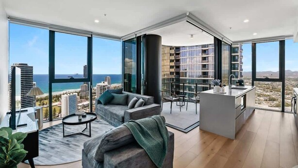 A modern living space with a large glass wall, a sofa, a dining area, and a kitchen. - Lvl 36 Ocean Views | 2BR skyhome | sky pool (Broadbeach)