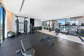 A modern gym with various exercise equipment, a bench, and a view of the city through large windows. - Lvl 36 at The Star | 2BR Ocean View, Pool, Parking (Broadbeach)