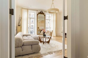 A living room with a sofa, a round coffee table, a mirror, and a view of the city through large windows. - The Primrose Princesa with A/C - by Out of Office Lifestyle (Greater London)