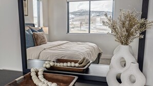A bedroom with a bed, a mirror, a vase with dried flowers, and a wooden tray with a string of white eggs.