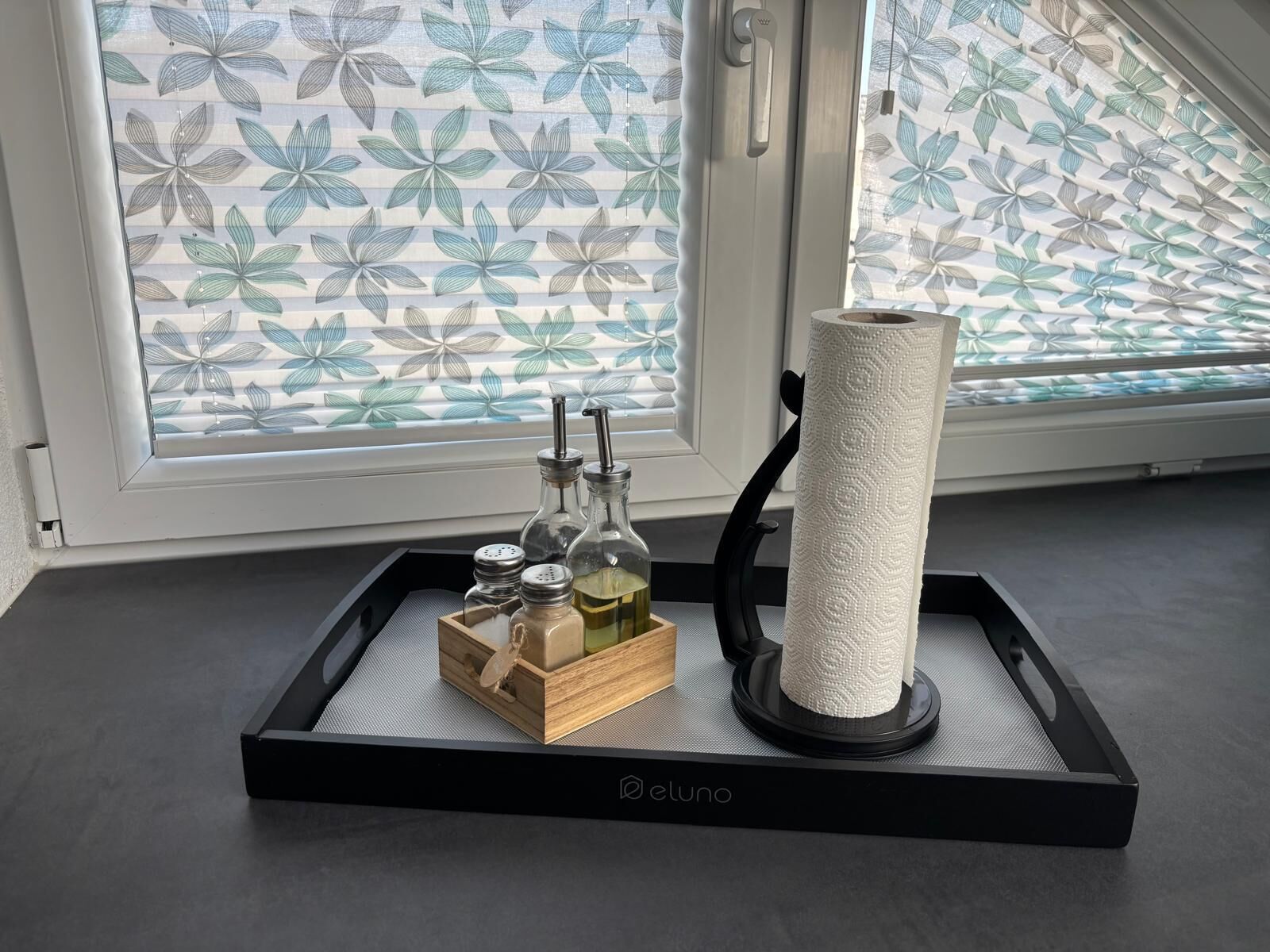 A tray with a tissue roll, salt and pepper shakers, and a small bottle on a black tray with a logo, placed on a windowsill with a floral blind.