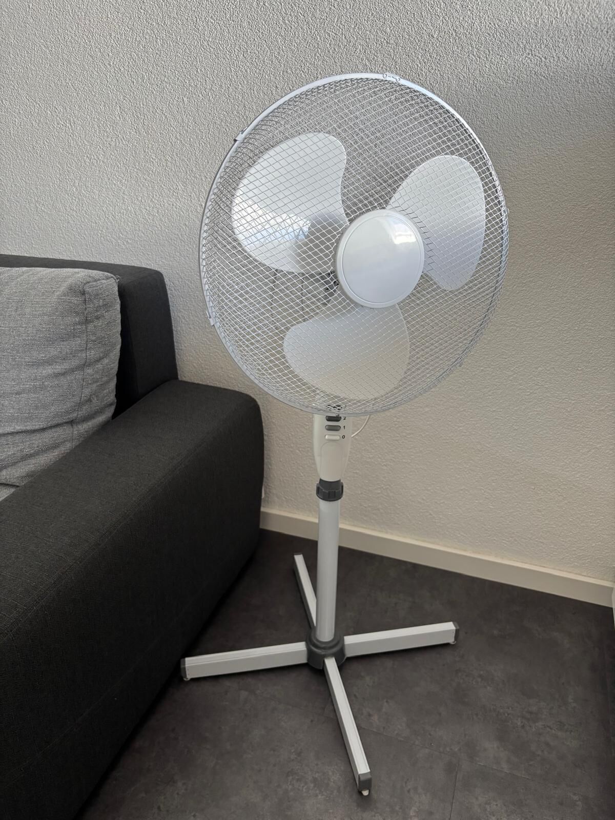 A standing floor fan with a mesh cover and a central light, positioned near a dark-colored sofa against a textured wall.