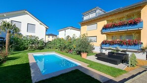 Zona residencial con piscina, césped cuidado y un edificio con balcones adornados con flores.