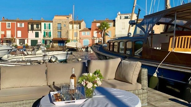 Mesa servida con una botella, vasos y flores en una terraza con vista a barcos y edificios de colores.