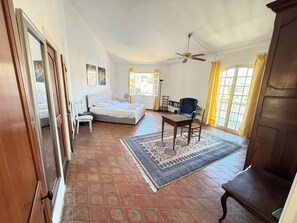 A bedroom with a bed, a desk, a chair, a ceiling fan, and a view through a window.