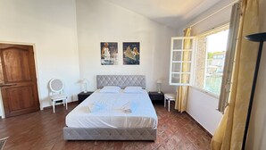 Un dormitorio con cama, una puerta de madera, una ventana con cortinas y dos cuadros colgados en la pared.