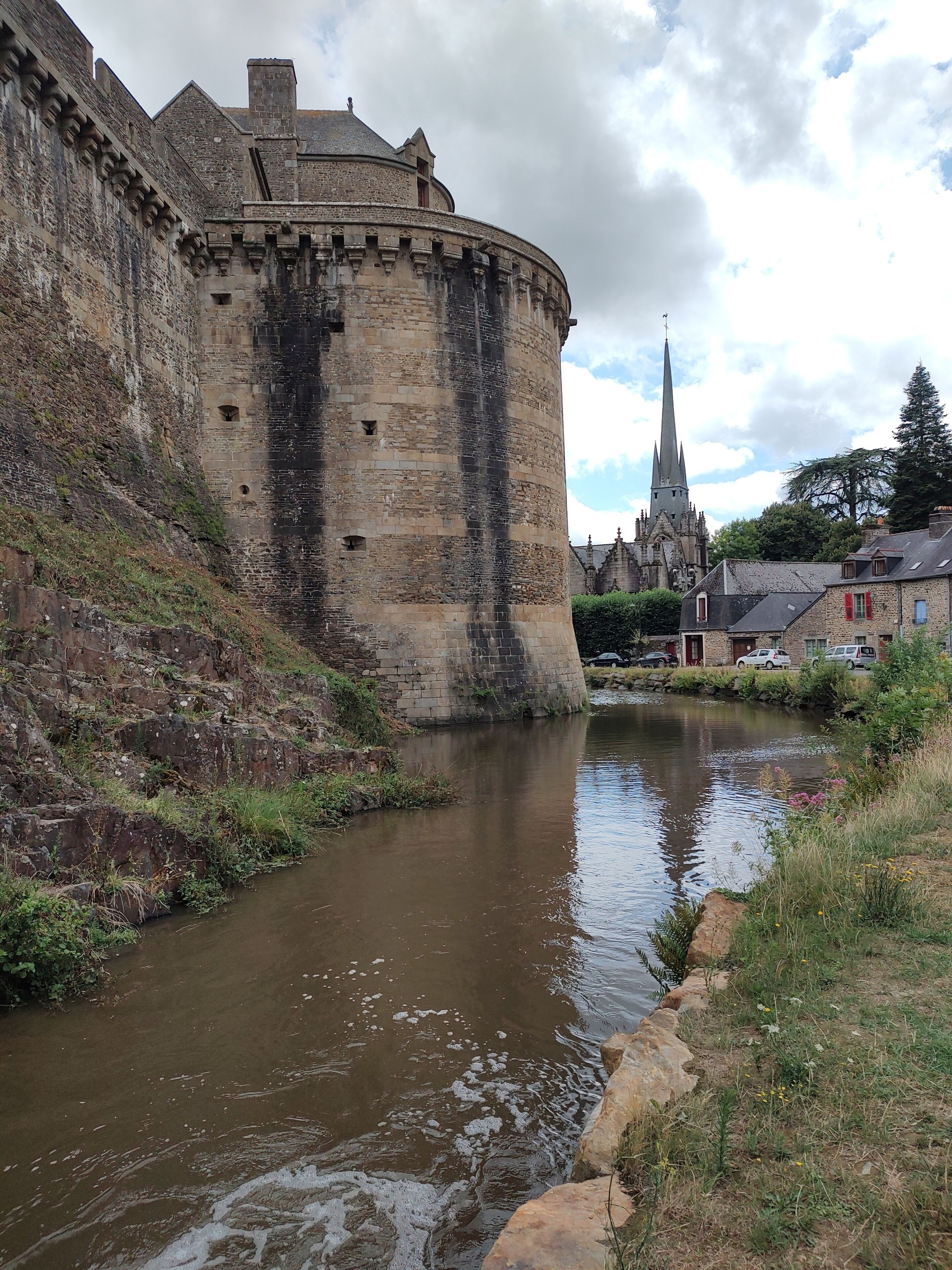 A historic stone castle with a moat, a church with a tall spire, and residential buildings with red shutters.