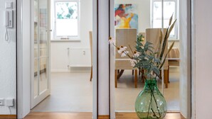 A room with a wooden floor, a green vase with dried plants, a white door, and a dining area with chairs and a table in the background. - Bächle Lodge with 3 Bedrooms and Mountain Views in the Heart of Hornberg (Hornberg)