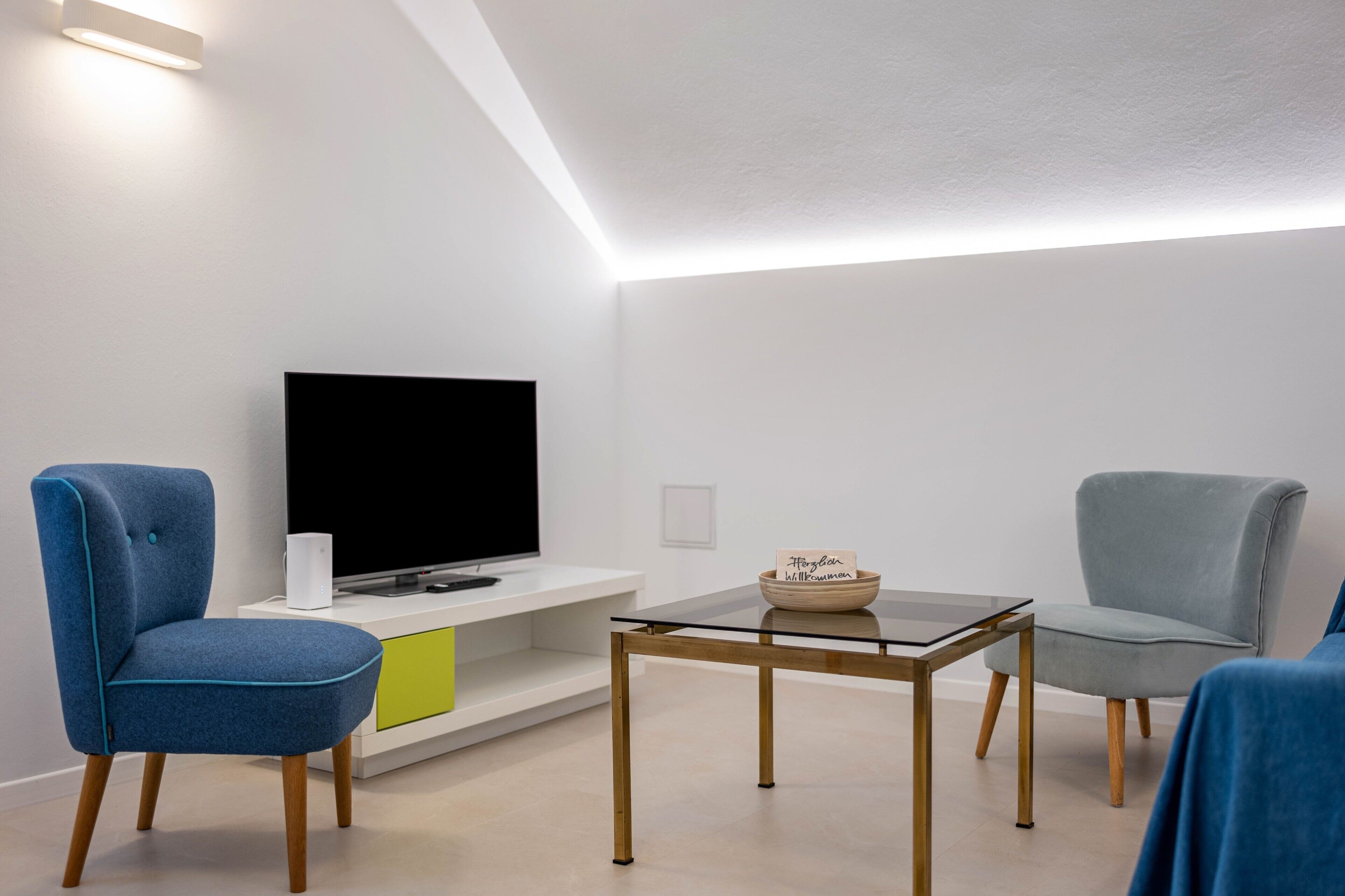 A modern living room with a flat-screen TV, two chairs, a glass coffee table, and a minimalist design.