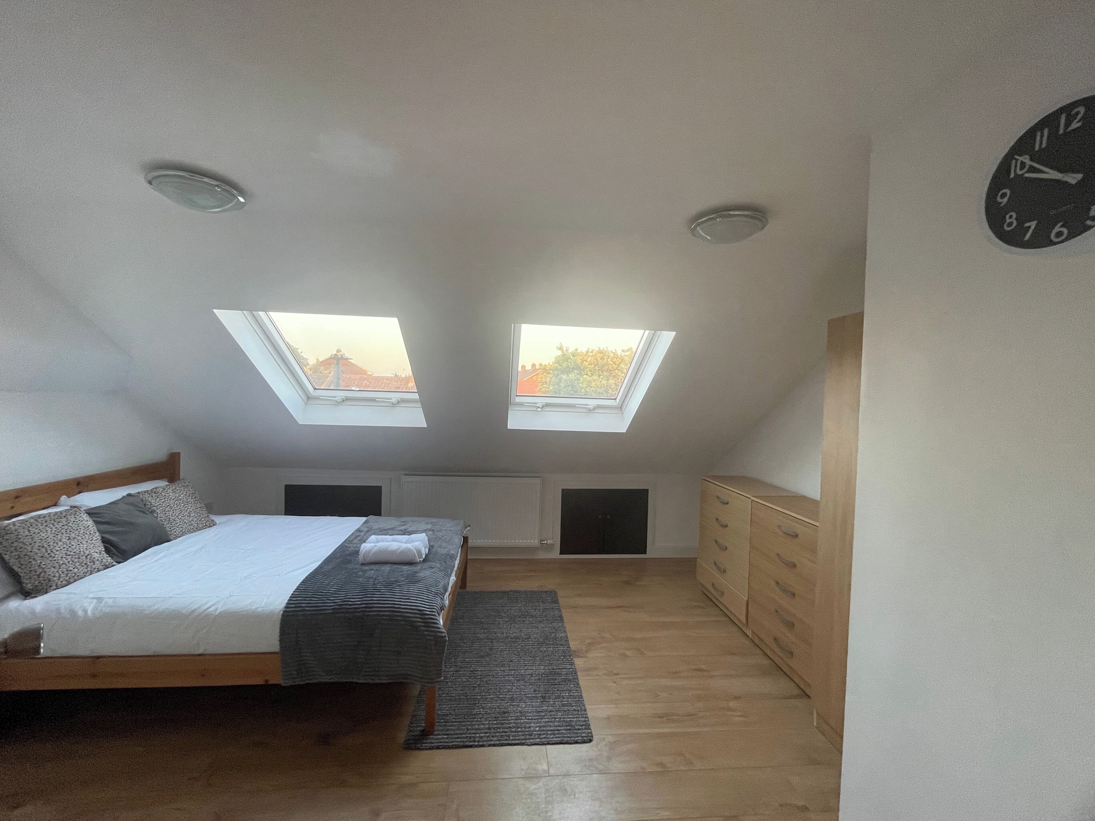 A bedroom with a skylight, a bed, a dresser, and a clock on the wall.