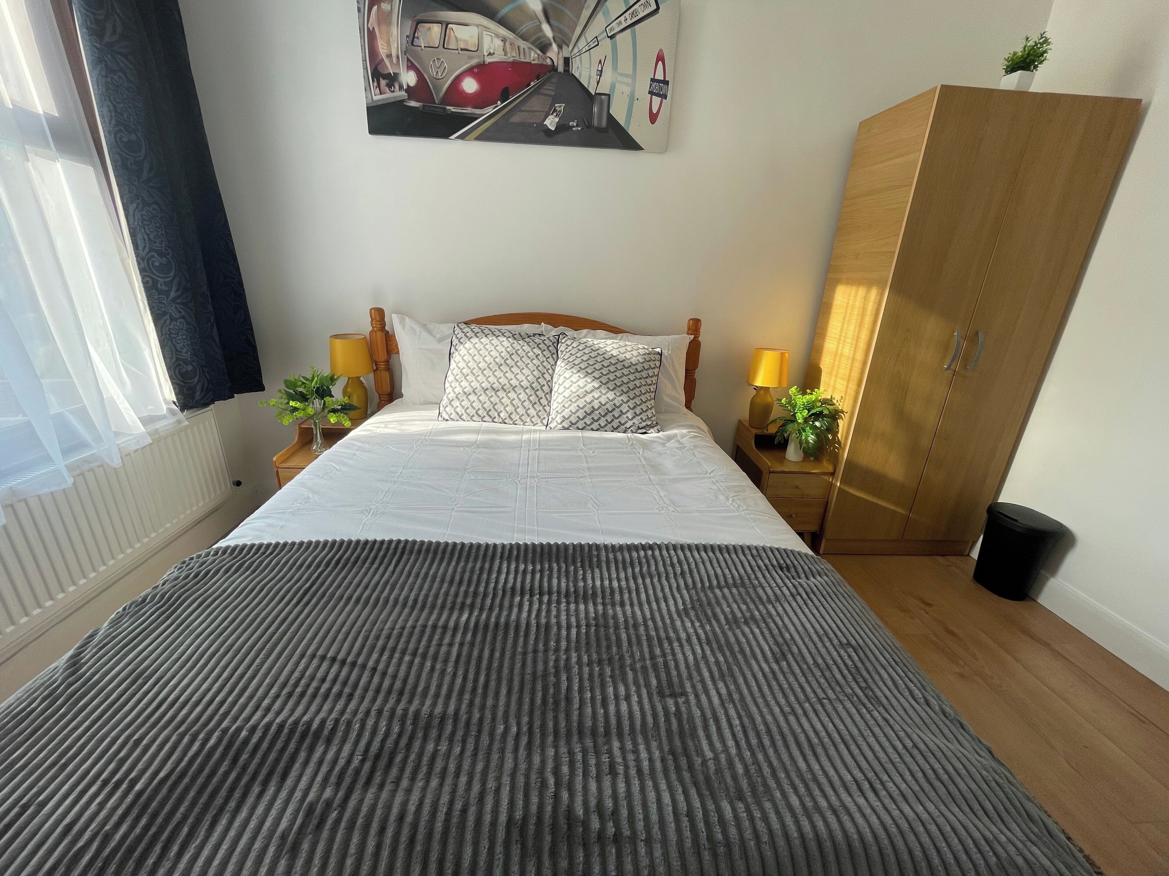 Ein ordentlich gemachtes Bett mit einer gestreiften Decke, zwei Nachttischlampen, einem Holzschrank und einem an der Wand hängenden Bild von einem roten Bus.
