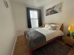 A bedroom with a wooden bed, a window with black curtains, a radiator, a wall clock, a painting of a train, and a bedside lamp.