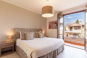 A bedroom with a large bed, a nightstand with a lamp, a window with a view of buildings, and a hanging light fixture. - Modern apartment 1, 5M Montepulciano with parking (Toscana)
