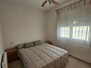 Une chambre à coucher avec un lit, un ventilateur de plafond, une fenêtre avec des rideaux et une petite table de chevet.
