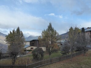 Um cenário rural com uma casa rodeada por árvores e montanhas ao fundo.