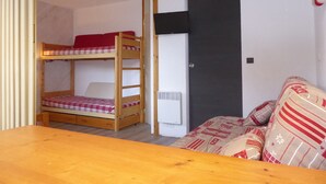 Un lit superposé avec un lit supérieur rouge, un lit simple en dessous, une petite table, un canapé et une porte avec un panneau noir.
