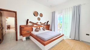 A bedroom with a wooden headboard, a bed with patterned pillows, a woven rug, and a view of the outdoors through a window with curtains. - Zanziland Apartments (Kiwengwa)