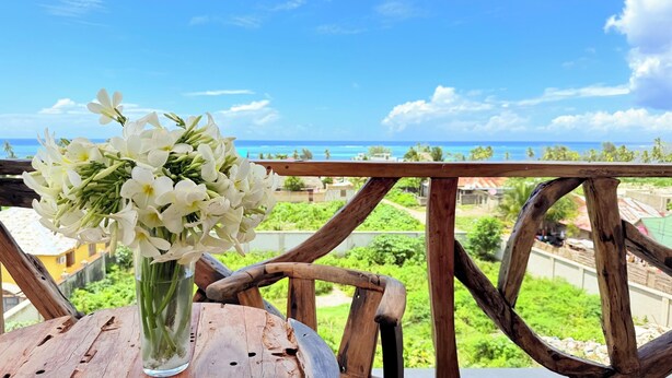 Une table en bois surmontée d’un vase de fleurs blanches, une balustrade en bois et une vue sur l’océan et la végétation.