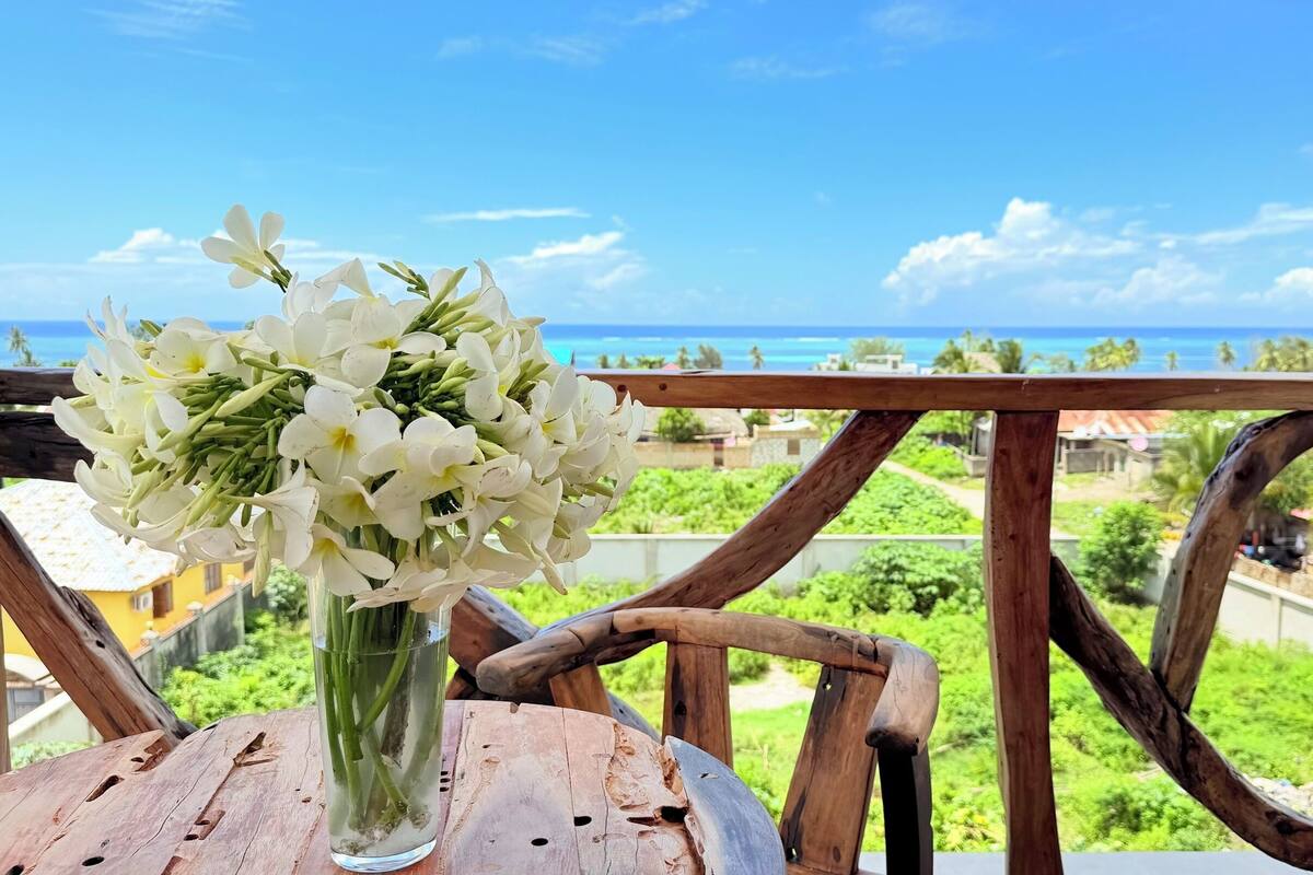 A wooden table with a vase of white flowers, a wooden railing, and a view of the ocean and greenery.
