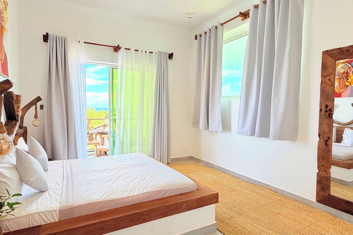 A bedroom with a bed, curtains, a large mirror, and a view of the outdoors.