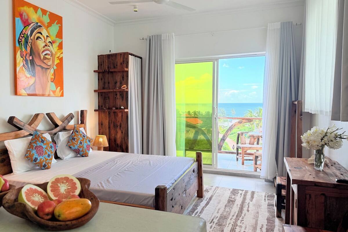 A bedroom with a bed, a wooden headboard, a nightstand, a fruit bowl, a painting, and a view of the outdoors.