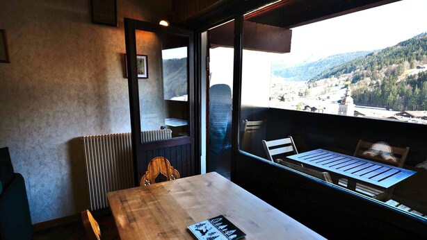 A dining area with a wooden table and chairs, a radiator, and a view of a mountainous landscape through a large window. - Studio with mezzanine near village center - 4 guests (Le Grand-Bornand)