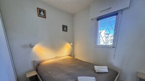 A single bed with a headboard, a small bedside table, two wall-mounted light fixtures, a window with a view of buildings, and a framed picture on the wall. - Apartment with sea view, parking - 4 guests (Six-Fours-les-Plages)