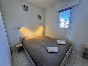 Een eenpersoonsbed met hoofdeinde, een klein nachtkastje, twee wandlampen, een raam met uitzicht op gebouwen en een lijst met een afbeelding aan de muur.