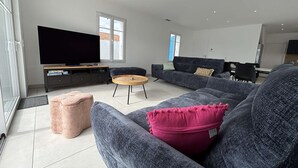 En moderne stue med et stort fladskærms-tv, grå sofaer, et træbord og en lyserød skammel.