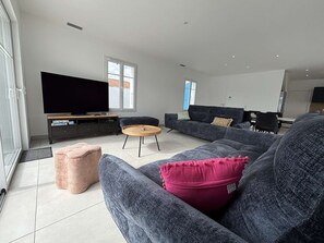 Una sala de estar moderna con un televisor de pantalla plana grande, sofás grises, una mesa de centro de madera y un reposapiés rosa.