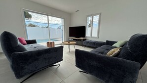 En moderne stue med to mørke sofaer, et rundt bord, et fladskærms-tv og et stort vindue med udsigt til en swimmingpool.