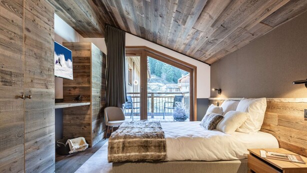 Ein Schlafzimmer mit Holzbalkendecke, einem Bett mit weißer Bettwäsche, einem Nachttisch aus Holz und Blick ins Freie.