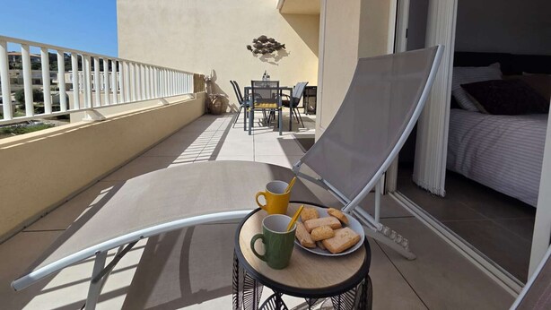 Un balcon avec une chaise, une petite table sur laquelle se trouve une assiette de biscuits et deux tasses, et une vue sur l’intérieur du bâtiment.