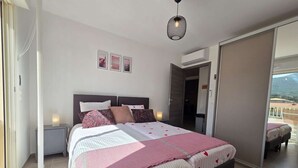 Une chambre à coucher avec un lit, des tables de chevet, un luminaire suspendu et une vue sur l’extérieur, visible à travers une porte ouverte.