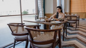 A dining area with a table set for two, wicker chairs, and a view of buildings through large windows. - Estrella The Resort (Karjat)