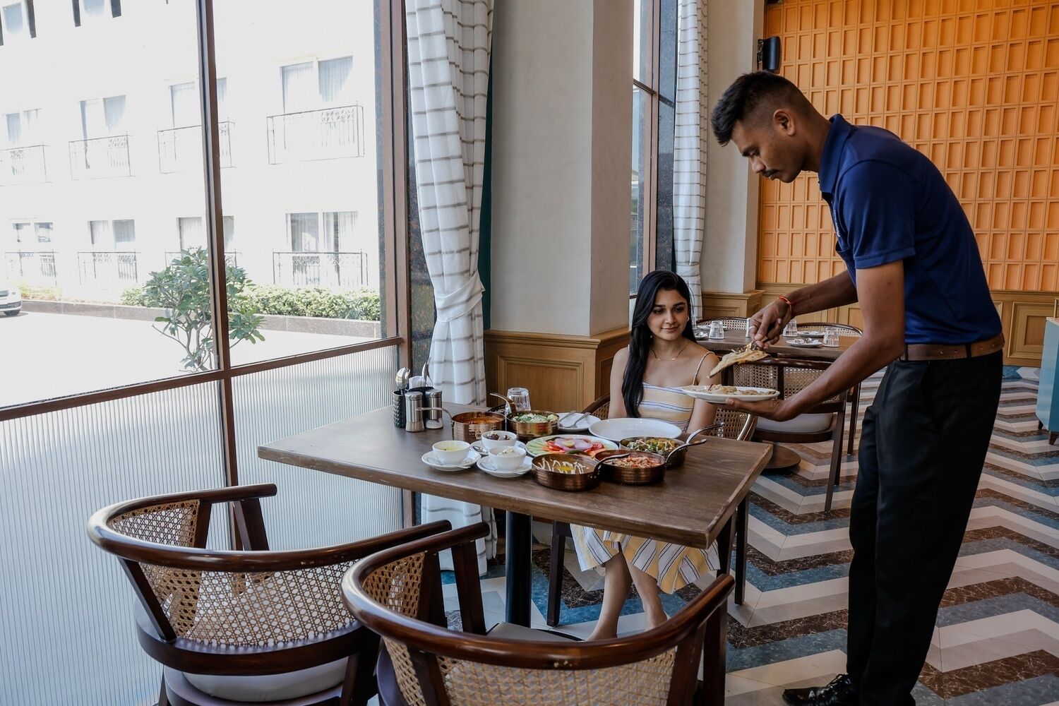 Un espace repas avec une table dressée pour deux personnes, un serveur qui apporte les plats et une vue sur un bâtiment à l’extérieur.
