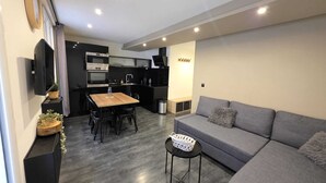 A modern living space with a grey sofa, a wooden dining table, a TV mounted on the wall, and a kitchen area with a built-in oven and microwave.