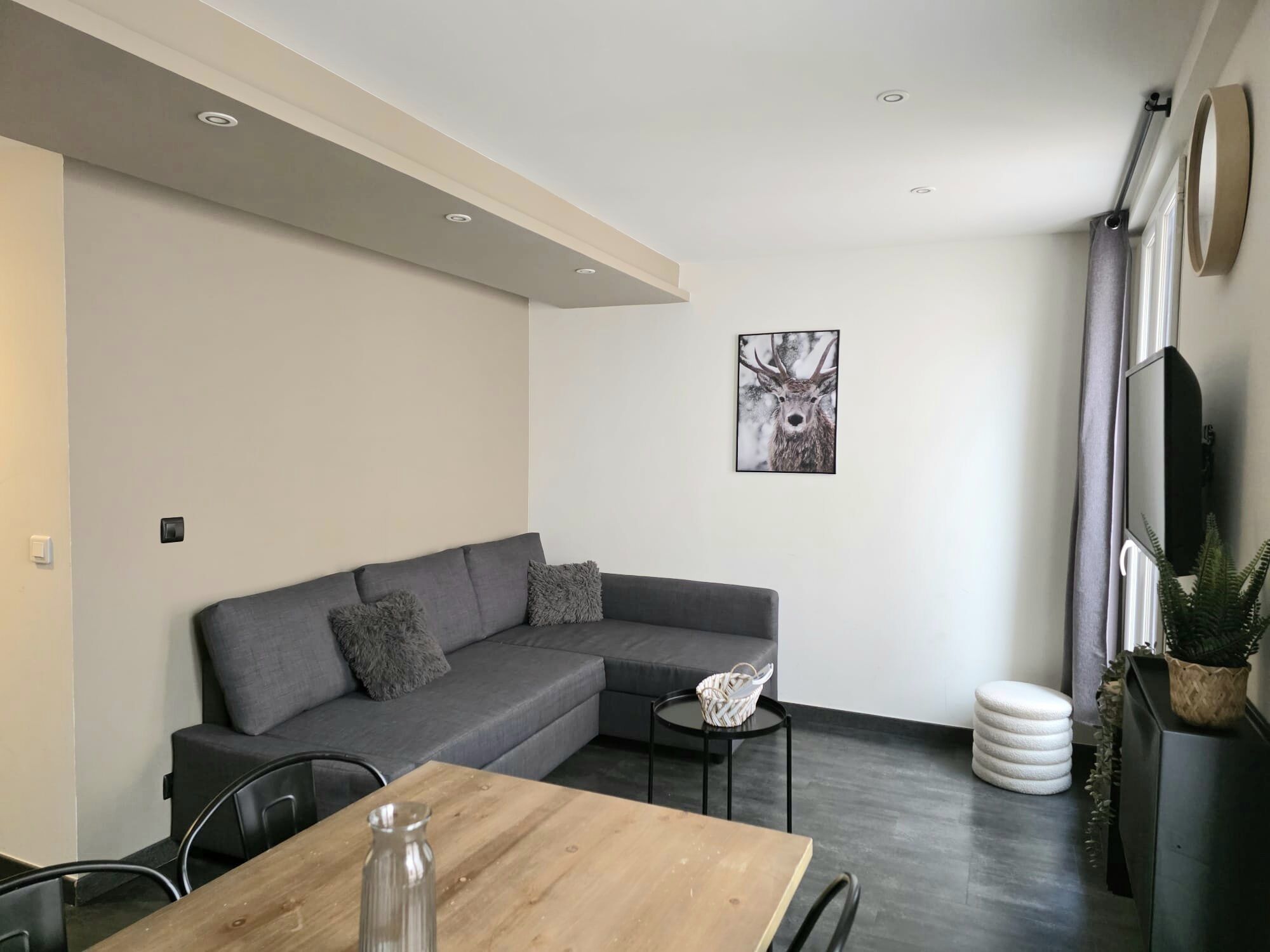 A modern living room with a grey sectional sofa, a wooden dining table, a small side table, a framed deer picture, a TV mounted on the wall, and a potted plant.