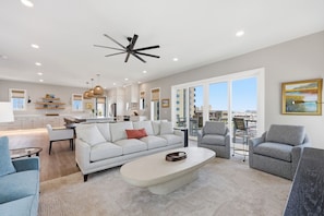 Ein geräumiges Wohnzimmer mit einer Couch, Sesseln und einem Couchtisch. Es gibt eine Küchenzeile mit einem Esstisch und Stühlen sowie einen Balkon mit Blick.