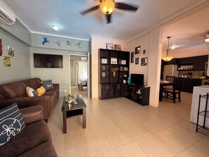 A living room with a brown sofa, a coffee table, and a TV stand. There is a kitchen area with cabinets and a refrigerator. - Tropical 2BR Condo in Playas del Coco – Pool, Walk to Beach & Downtown. (Playas del Coco)