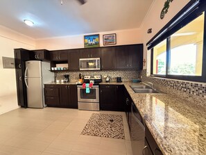 A modern kitchen with dark cabinets, stainless steel appliances, a tiled backsplash, and a window with a view. - Tropical 2BR Condo in Playas del Coco – Pool, Walk to Beach & Downtown. (Playas del Coco)