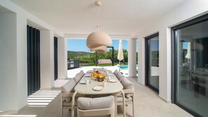 A modern dining area with a round table, cushioned seating, and a view of a swimming pool and greenery.