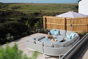 Ein gebogenes graues Sofa mit Kissen, ein kleiner Tisch mit Korb und Glas sowie ein Holzzaun in einem Garten.