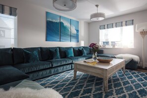 A living room with a dark blue sofa, a patterned rug, a coffee table with a bowl and flowers, and a large wave painting on the wall. - Polview (Newquay)