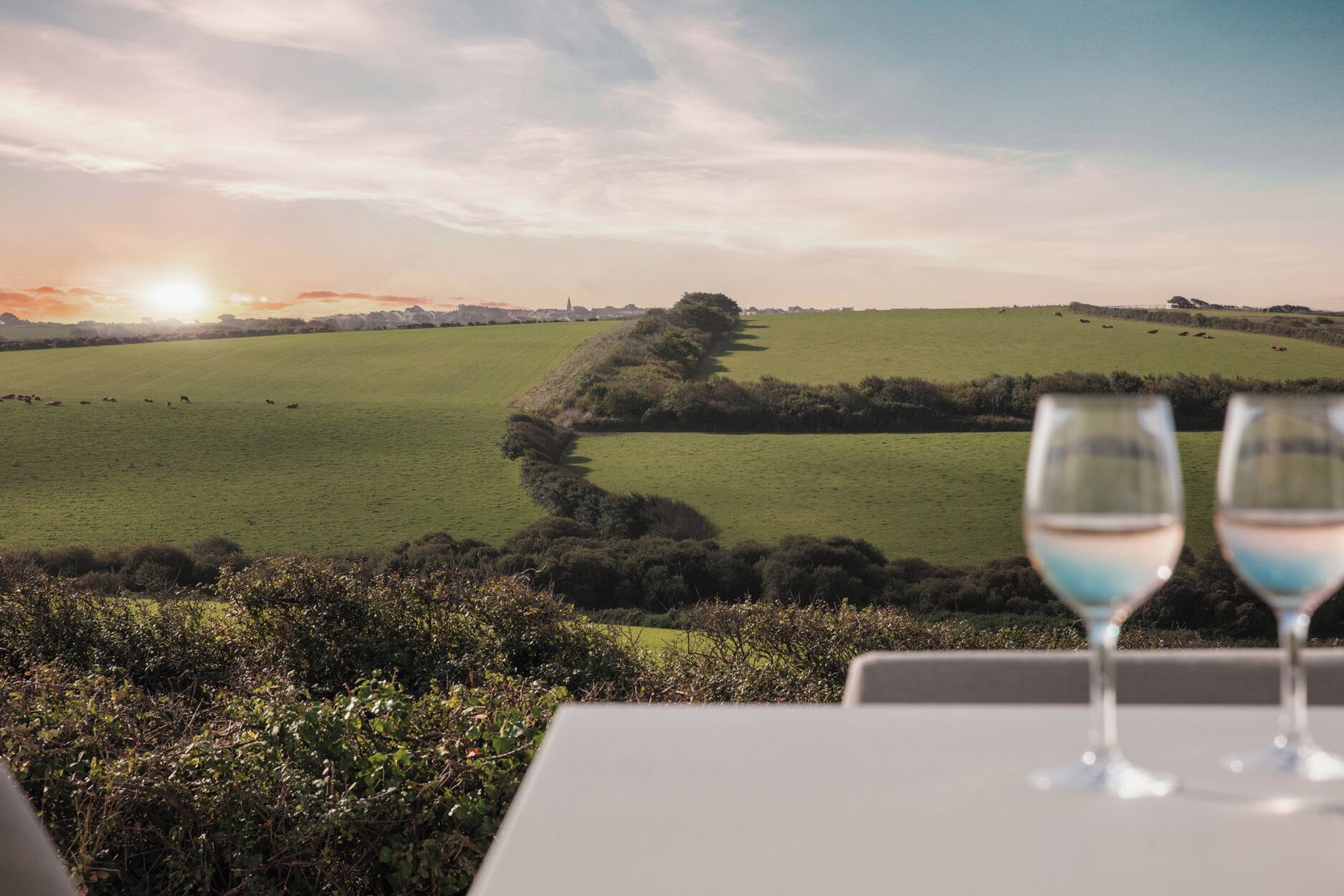 Ein Tisch mit zwei Weingläsern, Blick auf ein grünes Feld und ein Sonnenuntergang im Hintergrund.