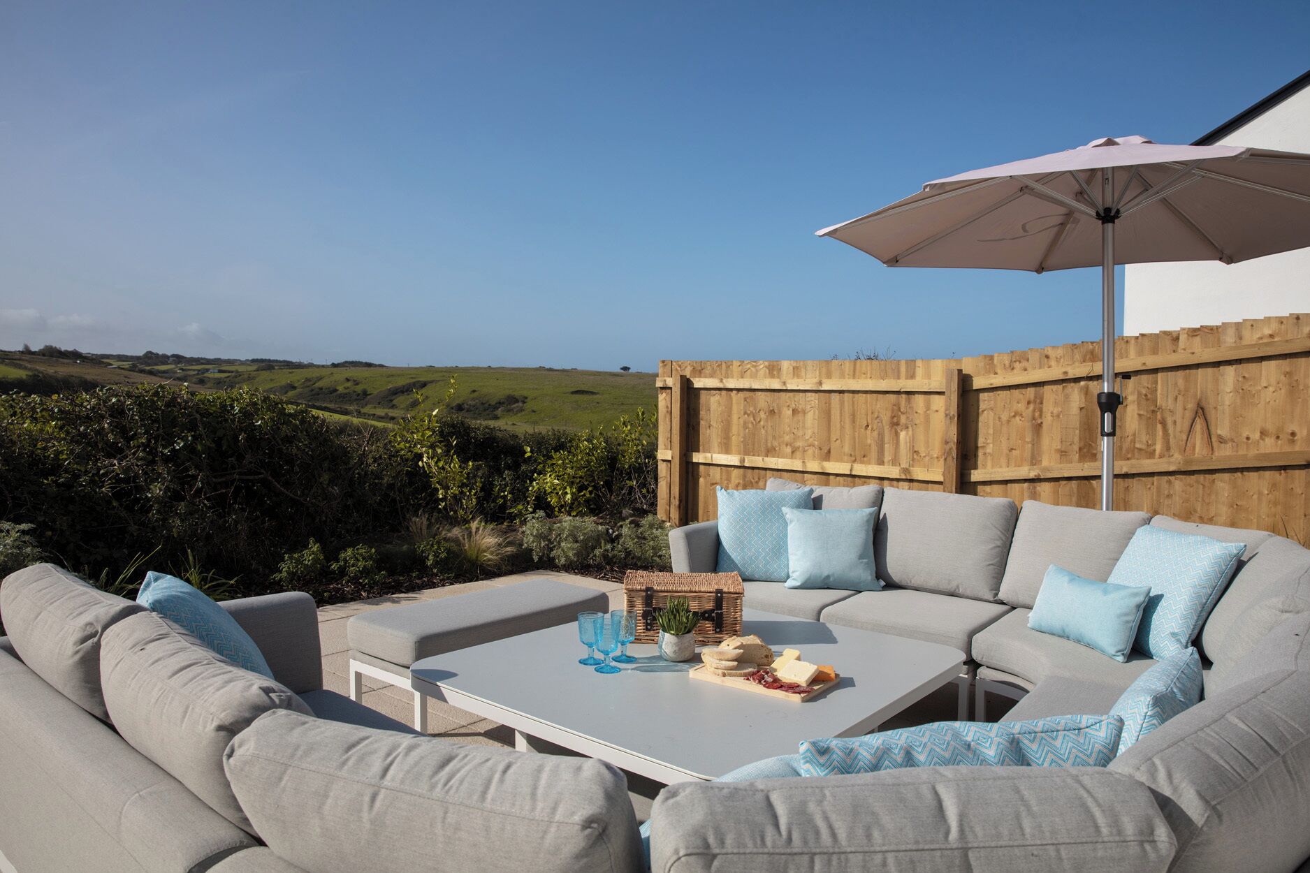 An outdoor seating area with a white table, grey sofa, and a wooden fence in the background.