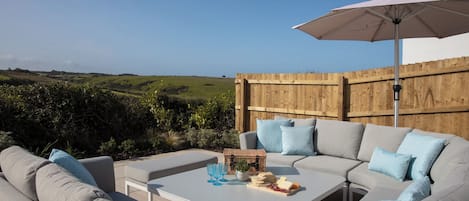 An outdoor seating area with a white table, grey sofa, and a wooden fence in the background.
