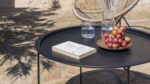 Un mur de pierre, une chaise en osier, une table ronde avec un livre et un bol de fruits, et une plante en pot.